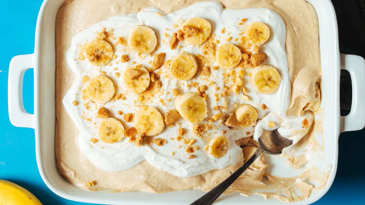 A rectangular dish holds a creamy dessert topped with whipped cream, banana slices, and crumbled cookies. A spoon rests inside, revealing a smooth, light brown peanut butter mixture underneath. A single banana is partially visible in the bottom left corner against a blue background. Satisfy your cravings with this treat!