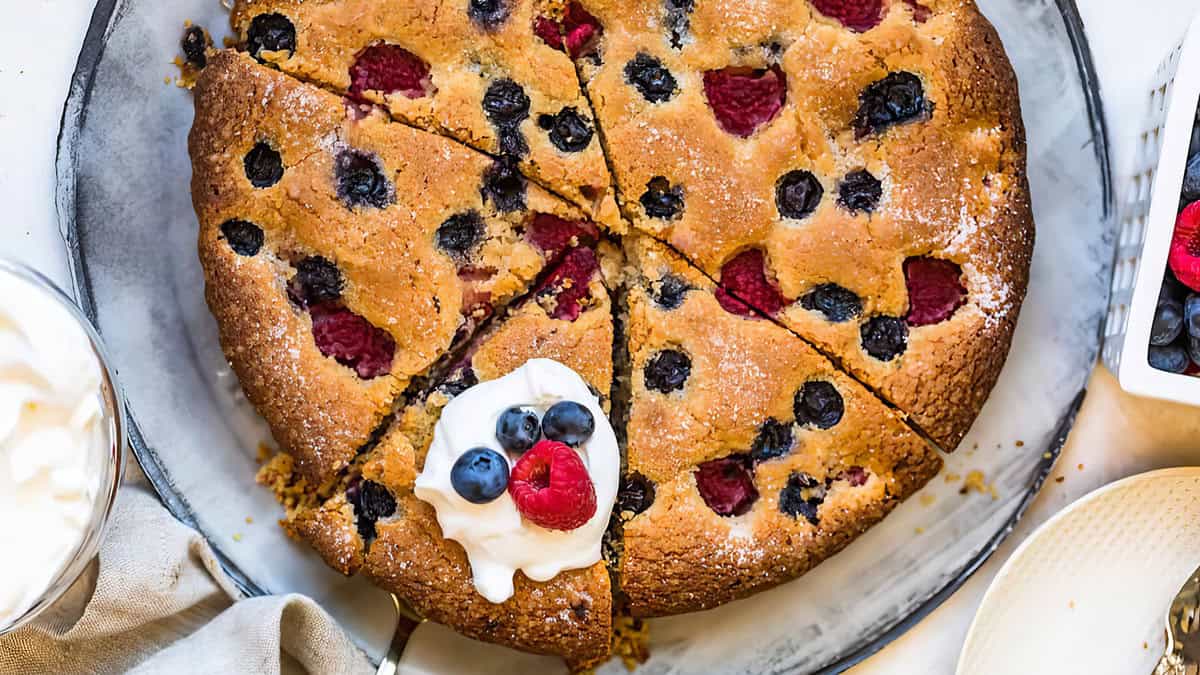 A freshly baked, irresistible coffee cake sliced into eight pieces. The golden brown cake is topped with blueberries and raspberries. One slice is garnished with whipped cream and extra berries. A bowl of whipped cream and more berries are visible in the background, perfect for a morning treat.