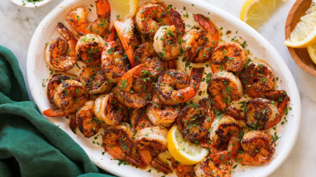 A white oval plate filled with seasoned, grilled shrimp garnished with chopped parsley and lemon wedges. A green napkin is partially visible on the left side, and additional lemon wedges are on the right. The background surface appears to be marble—a true delight from Southern recipes.