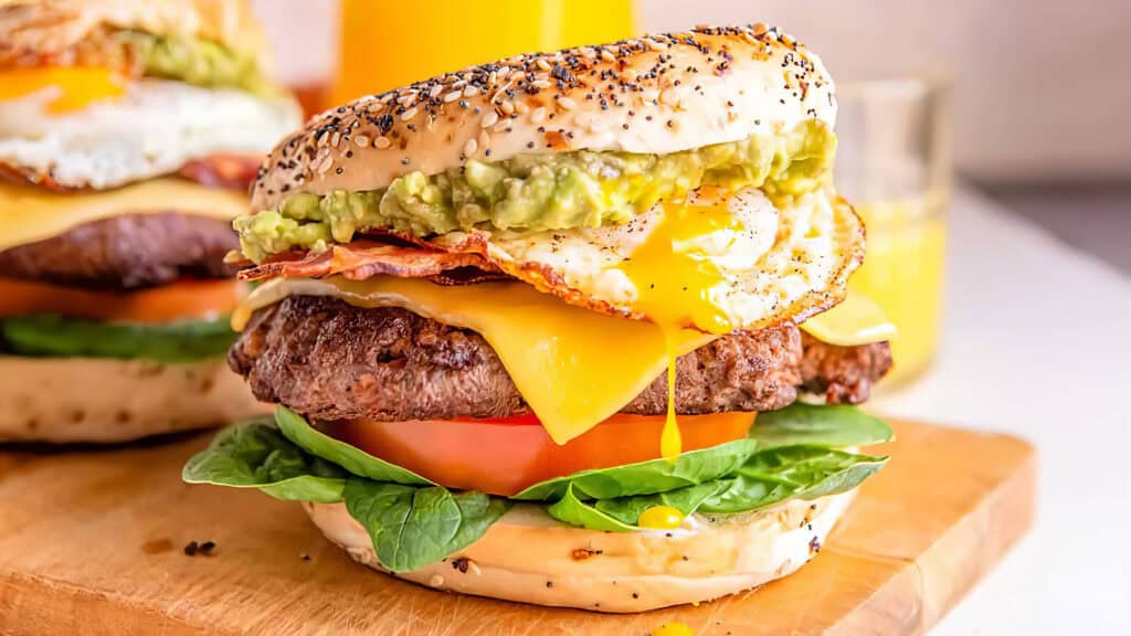 Close-up of a gourmet burger with a poppy seed bagel bun. The burger features layers of fresh spinach, thick beef patty, tomato slice, cheddar cheese, crispy bacon, a fried egg with runny yolk, and guacamole—worthy of pancake recipes. A glass of orange juice is in the background.