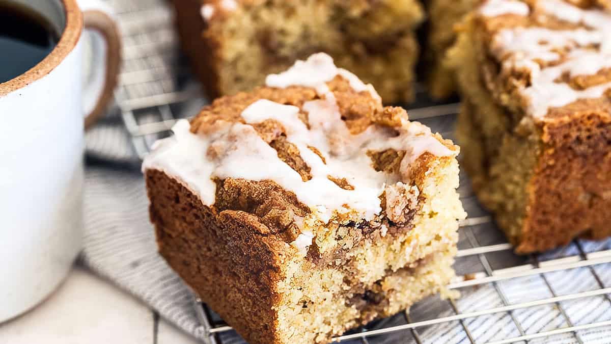 A close-up of a slice of irresistible crumb cake with a glaze drizzle, resting on a wire cooling rack. The slice shows layers of cake and a crumbly topping. A mug of black coffee sits beside the cake, partially visible on the left—wake up and smell the coffee cake!