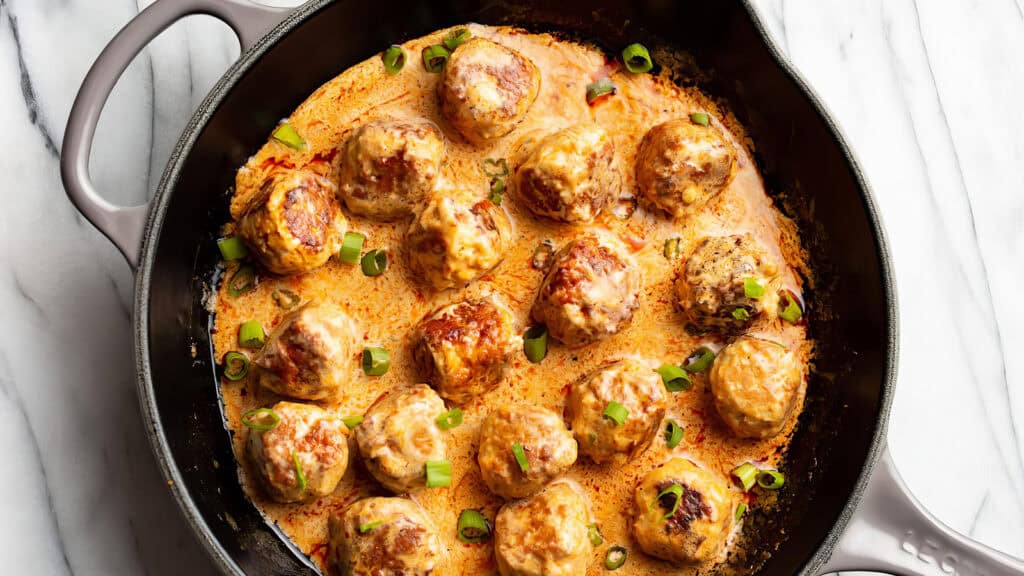 A black cast-iron skillet filled with creamy sauce and round meatballs garnished with chopped green onions rests on a white marble surface. Perfect for any cookbook, the meatballs are browned and cooked through, covered in a rich, orange-colored sauce—a must-try recipe!