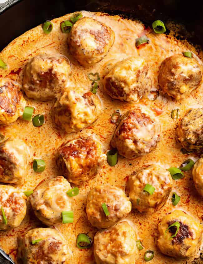 A black cast-iron skillet filled with creamy sauce and round meatballs garnished with chopped green onions rests on a white marble surface. Perfect for any cookbook, the meatballs are browned and cooked through, covered in a rich, orange-colored sauce—a must-try recipe!