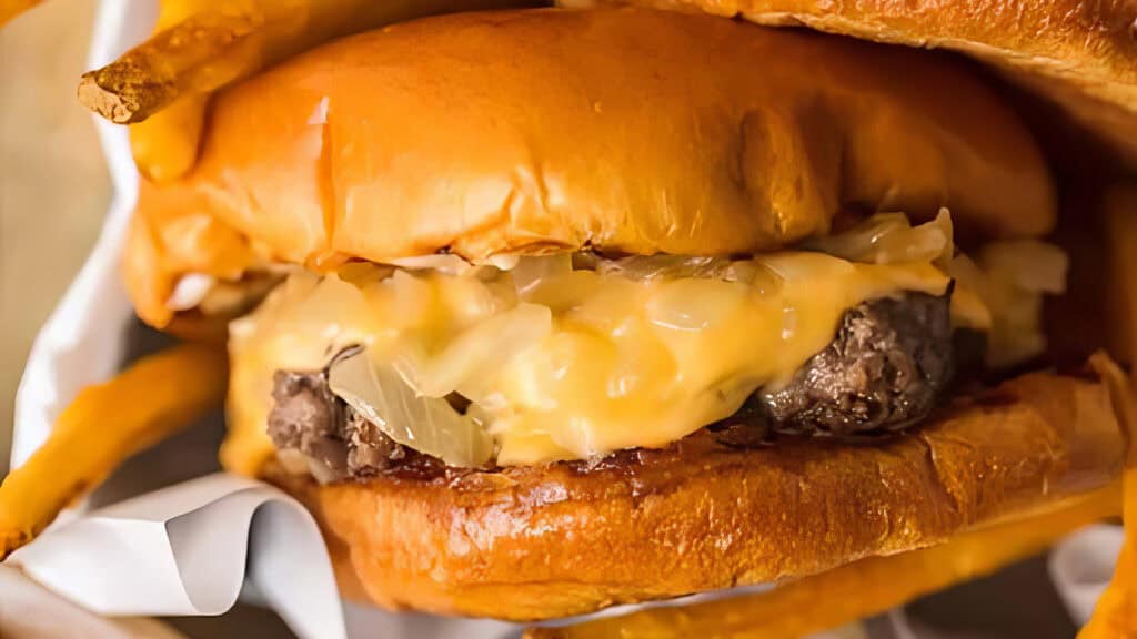 A close-up view of a cheeseburger with melted cheese, caramelized onions, and a juicy beef patty crafted from ground beef recipes, all nestled between toasted buns. Some French fries are visible in the background, adding to the appetizing appearance of the meal.