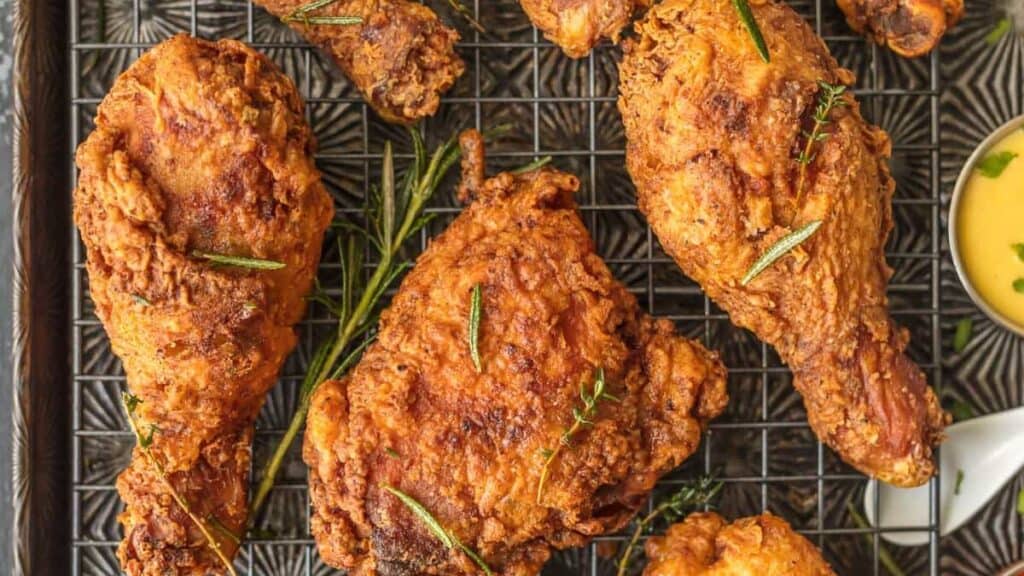 A tray of crispy fried chicken pieces, inspired by Southern recipes, is garnished with herbs. The chicken is arranged on a wire rack with a patterned tray beneath. A small dish of yellow dipping sauce is visible on the right side. Both drumsticks and other cuts are present.