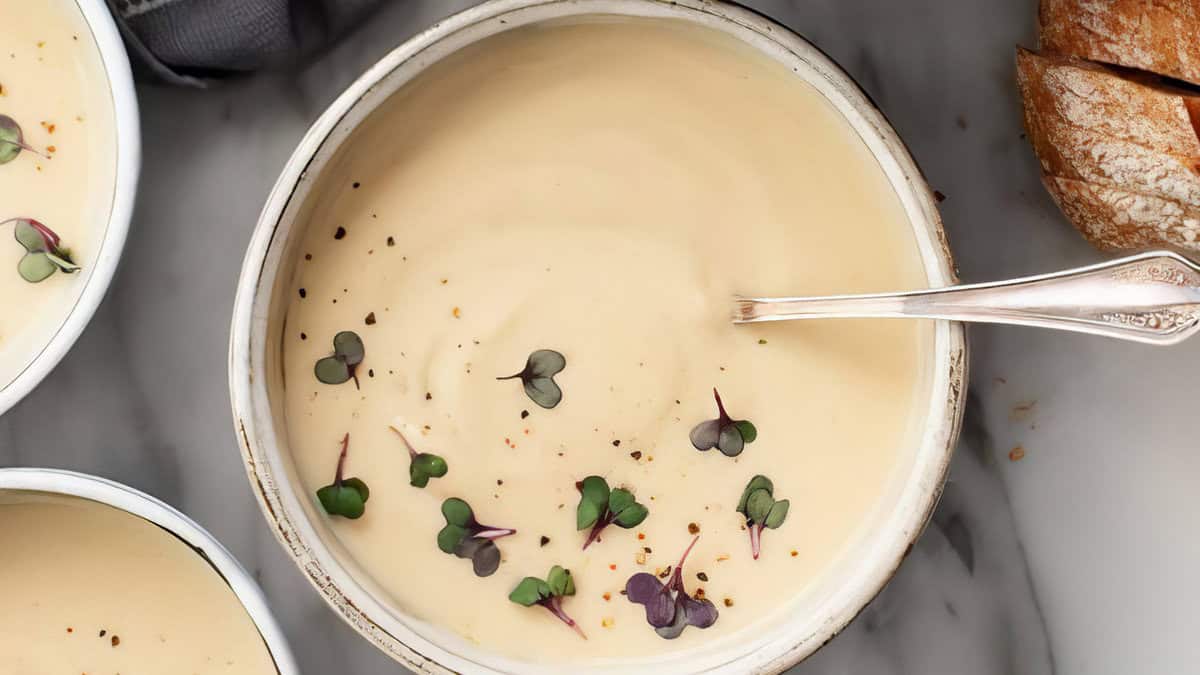 Close-up of a bowl of creamy comfort food soup garnished with microgreens and sprinkled with black pepper. A silver spoon rests in the bowl. In the background, there is a partial view of sliced crusty bread on the right and a smaller bowl on the left, all on a marble surface. Warm Your Soul.