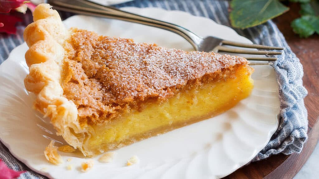 A slice of custard pie with a golden-brown crust is served on a white plate. The pie, reminiscent of classic Southern recipes, has a lightly browned, crumbly topping and is dusted with powdered sugar. A fork rests next to the slice on the plate, which sits on a gray cloth with green leaves in the background.