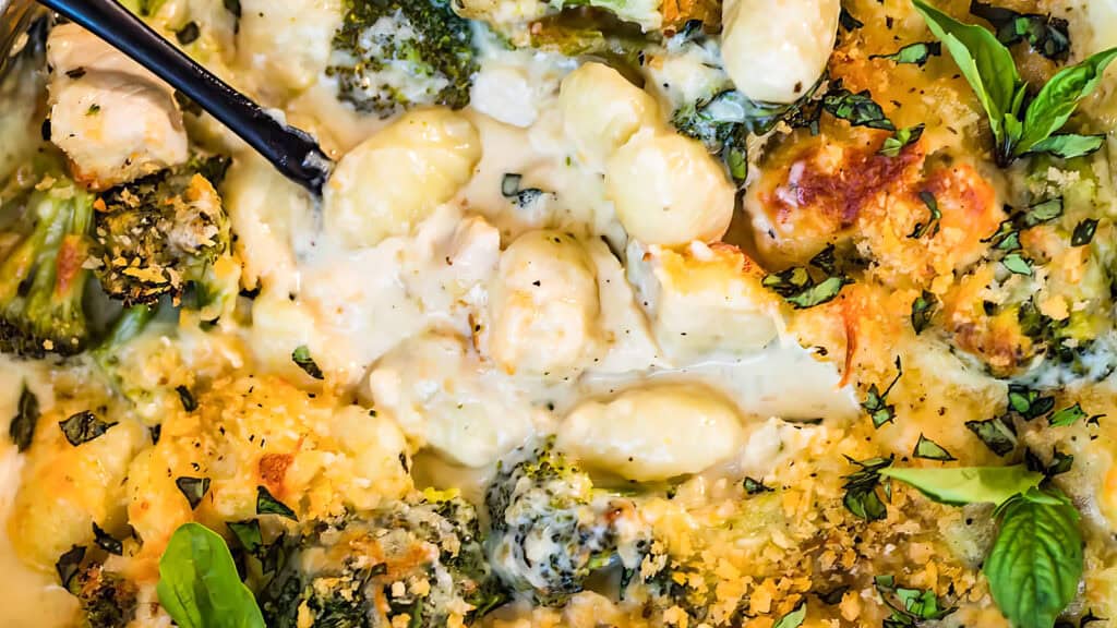 Close-up of a creamy baked gnocchi dish with broccoli florets and a golden breadcrumb topping, garnished with fresh herbs and basil leaves. Inspired by good Alfredo recipes, this savory meal is ready to be served with the black utensil partially visible in the upper-left corner.