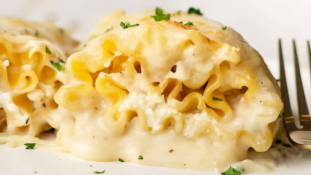 A close-up of creamy, cheesy lasagna roll-ups served on a white plate. The filling is visible, consisting of ricotta cheese, smothered in a rich Alfredo sauce and garnished with chopped parsley. A fork rests beside the lasagna roll-ups, making it look so good you'll forget about the calories.