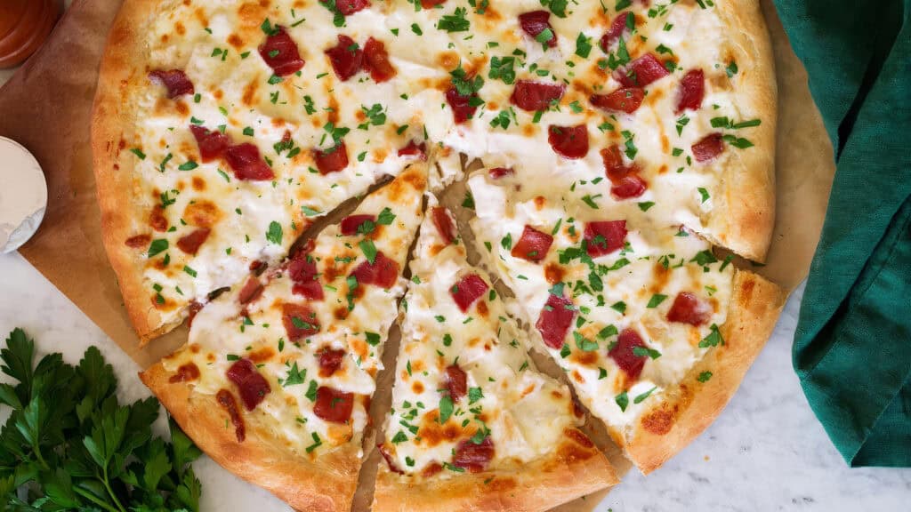 A close-up view of a freshly baked pizza topped with melted cheese, bacon bits, and chopped parsley. One slice has been cut and slightly pulled away from the rest of the pizza, revealing the golden-brown crust and creamy white Alfredo sauce underneath—a true delight for fans of rich Alfredo recipes.