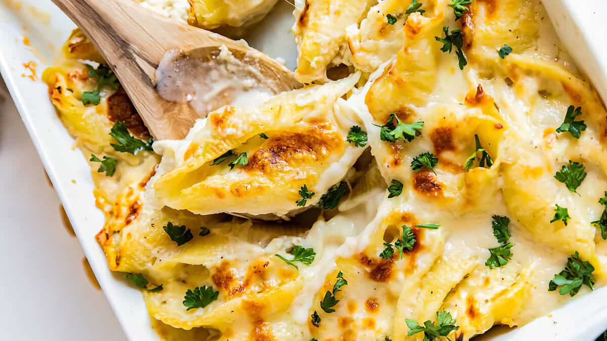 Close-up of a creamy baked pasta dish garnished with chopped parsley in a white baking dish. A wooden spoon is scooping a portion, revealing a gooey, cheesy texture with browned, crispy edges—this is one of those Alfredo recipes that's worth every calorie.