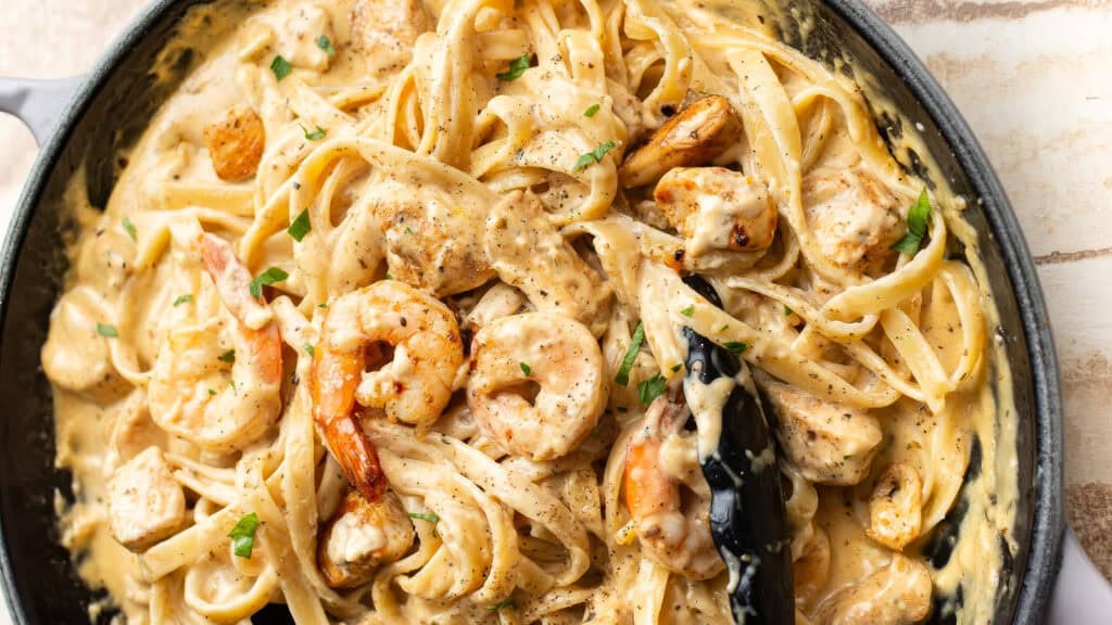 A close-up of creamy Cajun Chicken and Shrimp Alfredo pasta in a black skillet. The dish features fettuccine noodles coated in a rich, creamy sauce with large shrimp, pieces of chicken, and sprinkled parsley. A pair of tongs is holding some pasta, ready to serve one of the best Alfredo recipes you'll find.