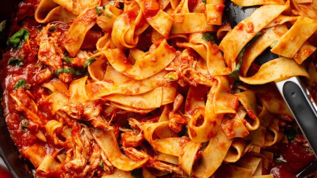 A close-up shot of a pan filled with pappardelle pasta mixed with a rich tomato sauce, reminiscent of Mediterranean recipes. Shredded chicken and fresh herbs are visible throughout the dish. A serving spoon is partially shown in the upper right corner, contrasting against the vibrant red sauce.