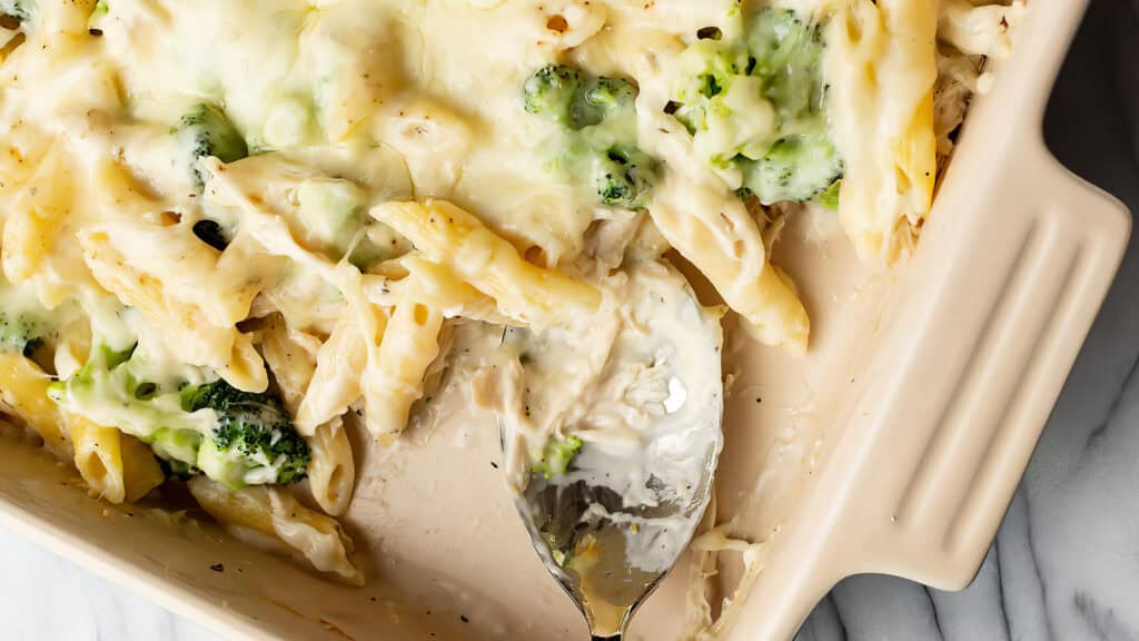 A close-up of a creamy baked pasta dish in a rectangular ceramic baking dish. The pasta, inspired by classic Alfredo recipes, is mixed with broccoli florets and covered in melted cheese. A silver spoon is partially immersed in the casserole, indicating it has been served from.