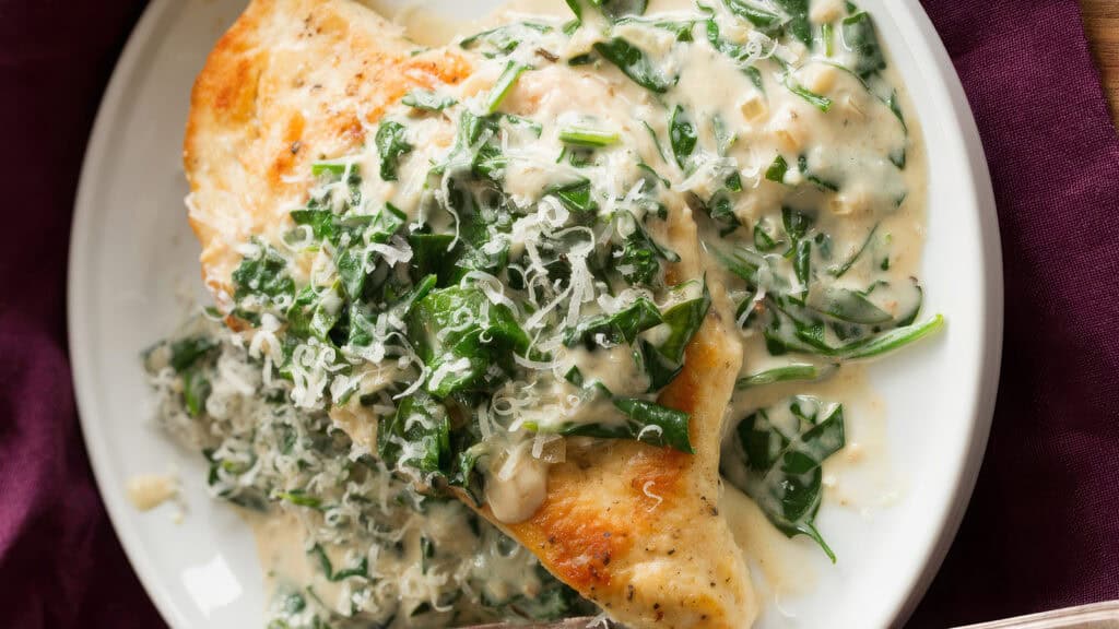 A plate of creamy garlic Parmesan chicken is pictured, showcasing one of the finest chicken recipes. Golden-brown chicken breasts are topped with a rich, creamy sauce and garnished with fresh spinach and grated Parmesan cheese. The dish is elegantly presented on a white plate with a burgundy napkin beside it.