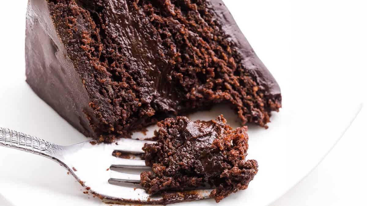 A close-up of a rich, moist slice of chocolate cake with thick, glossy chocolate frosting on a white plate. A fork, holding a bite-sized piece of cake made with avocado for a healthier twist, is placed beside the slice, showing the dense and fudgy texture of the dessert.