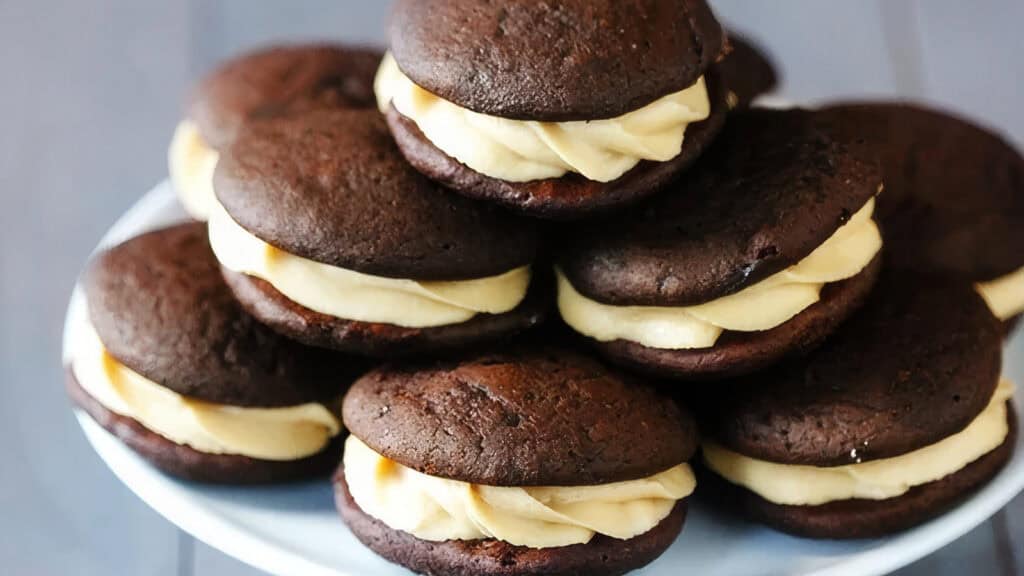 A plate stacked with chocolate whoopie pies filled with creamy vanilla frosting. The whoopie pies, part of irresistible chocolate recipes, are rich brown in color and arranged in a pyramid style, showcasing their thick filling and soft, cake-like texture.