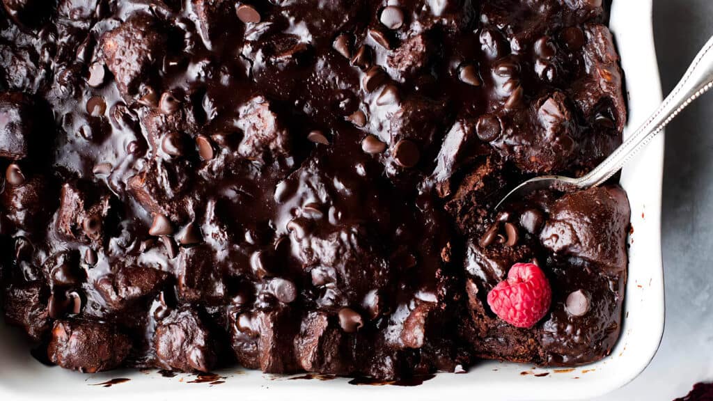 A close-up of a rich, gooey chocolate bread pudding from delectable chocolate recipes, topped with melted chocolate chips in a white baking dish. A spoon is scooping a portion, revealing a fresh raspberry nestled on the edge of the pudding.