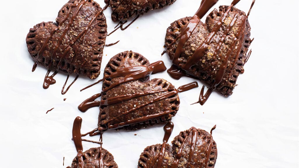 Five heart-shaped chocolate pastries with crimped edges are arranged on a white surface. Drizzled with melted chocolate and sprinkled with glittery sugar, these delightful sweets represent the pinnacle of Chocolate Recipes, creating a visually appealing, sweet treat.