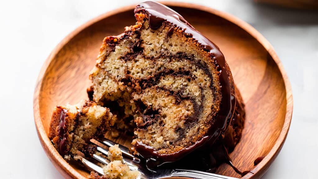 A close-up of a slice of marble cake with chocolate and vanilla swirls, sitting on a wooden plate. The cake is drizzled with rich chocolate sauce and a fork rests beside it, partially embedded in the cake—a true delight for lovers of Bundt Cake Recipes.