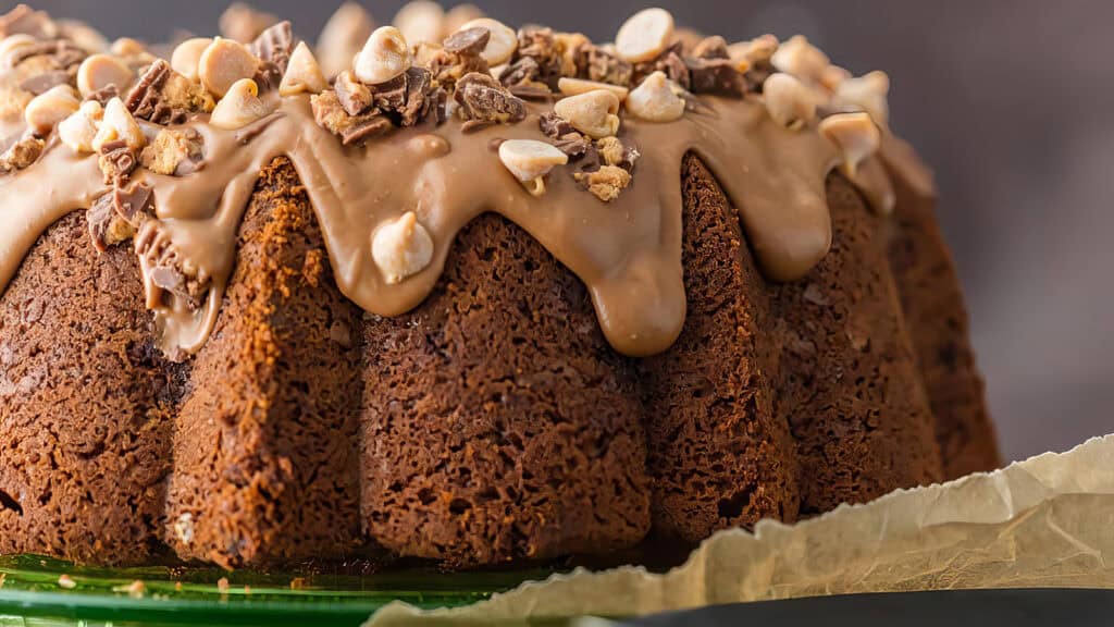 A close-up of a chocolate Bundt cake topped with a smooth chocolate glaze and sprinkled with chopped nuts and chocolate pieces, perfect for your Bundt Cake Recipes collection. The cake is on a green plate with parchment paper peeking from underneath. The background is blurred.