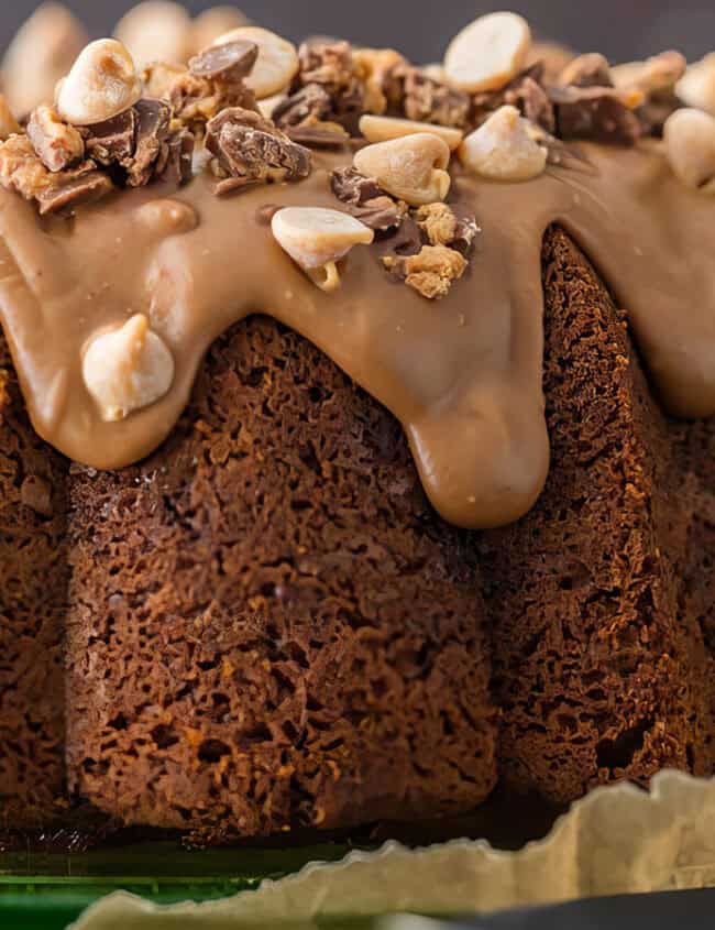 A close-up of a chocolate Bundt cake topped with a smooth chocolate glaze and sprinkled with chopped nuts and chocolate pieces, perfect for your Bundt Cake Recipes collection. The cake is on a green plate with parchment paper peeking from underneath. The background is blurred.