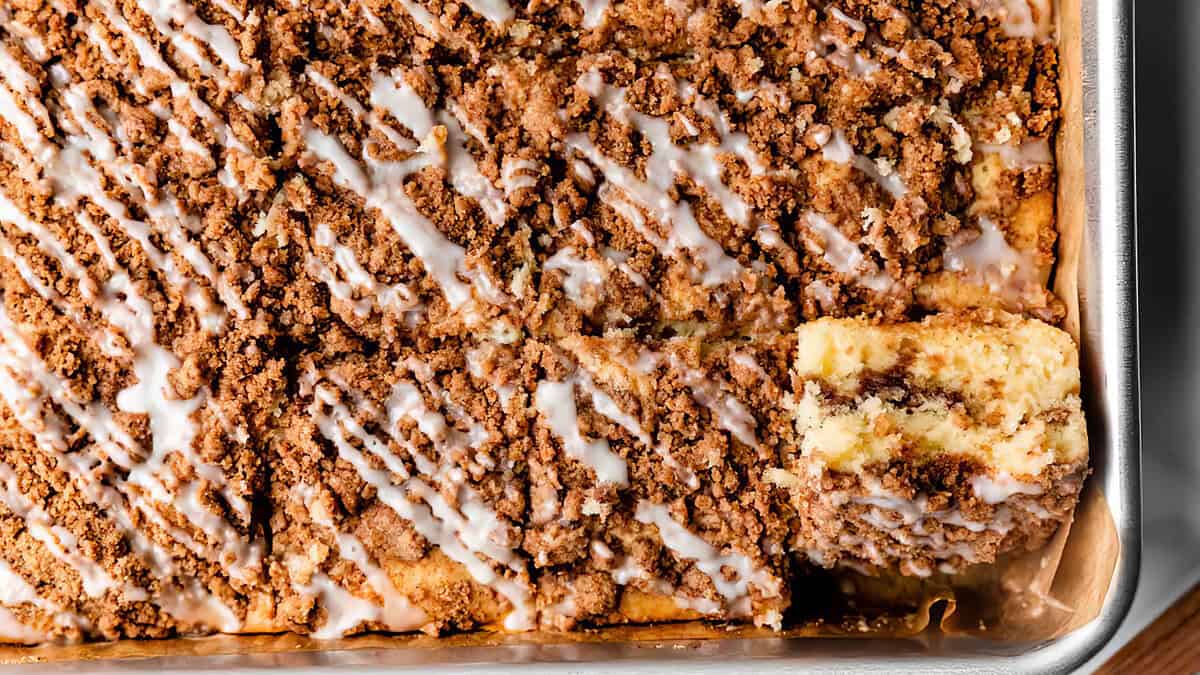 A close-up of a rectangular crumb cake with cinnamon streusel topping and white icing drizzle in a metal pan. One corner piece is cut and slightly pulled out, revealing the moist interior and layers of cake and streusel. Wake Up and Smell the Coffee Cake with these irresistible recipes!