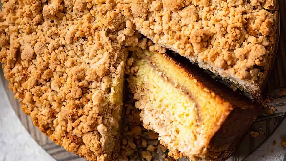 A close-up of a crumbly coffee cake with a slice being slightly removed to show its moist, layered interior. The cake has a golden-brown crumble topping, and the slice reveals a light, creamy layer nestled between soft, cake layers—one of those irresistible recipes you can't pass up.