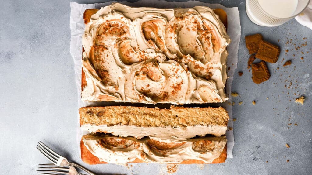 A rectangular cake topped with swirled brown frosting is cut into two halves. Crushed cookies are sprinkled on the frosting. A whole cookie, crumbs, and a partially full glass of milk are beside the cake on a gray background, with two forks placed near the cake—a perfect addition to your sheet cake recipes.