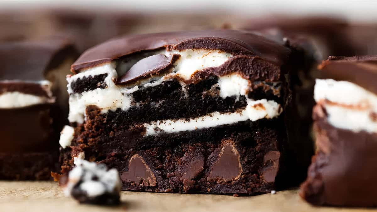 A close-up of a rich, layered dessert bar perfect for chocoholics, featuring a chocolate base with chocolate chunks, a white crème layer, and a thick, glossy dark chocolate topping. A bite has been taken out of the front piece, revealing its brownie-like texture.