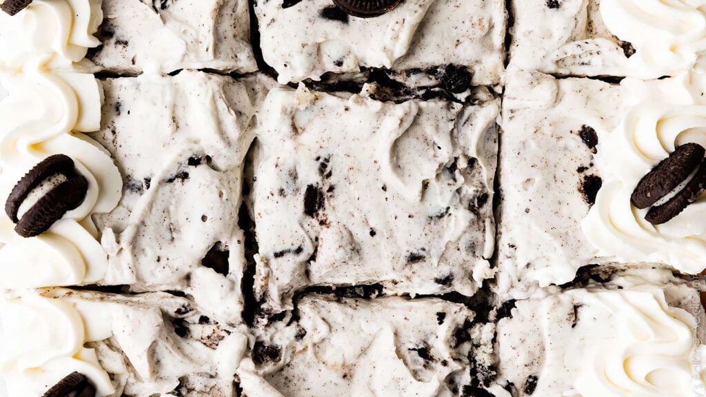 Top view of a batch of cookies and cream ice cream bars cut into squares. The bars feature a visibly creamy texture with chunks of cookies. Echoing popular sheet cake recipes, the edges are adorned with swirls of whipped cream and mini chocolate sandwich cookies as decoration.