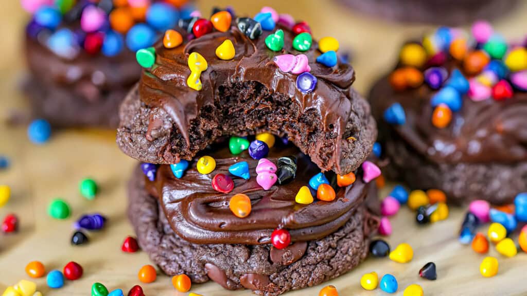 Close-up of chocolate cookies topped with chocolate frosting and colorful candy-coated pieces. One cookie, with a bite taken out, is stacked on another. More cookies and candy pieces are scattered in the background, showcasing irresistible treats from our best chocolate recipes.