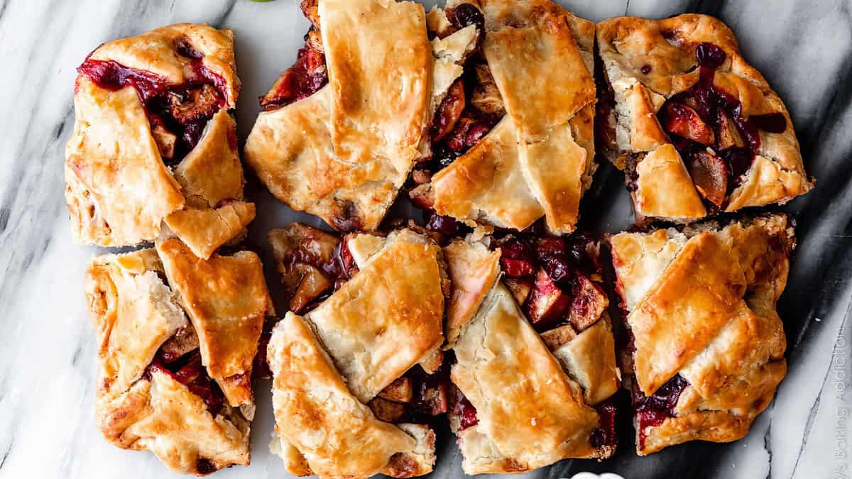 A close-up of freshly baked dessert pie squares on a marble surface. The pie squares have a golden brown, flaky crust with visible patches of baked fruit filling, including pieces of various fruits peeking through the gaps. The fruit juices have lightly caramelized, perfect for sweet recipes.