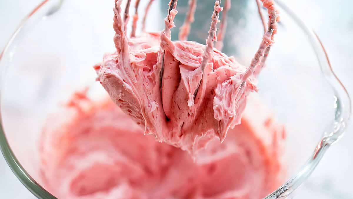 A close-up view of pink frosting being whipped by a mixer. The fluffy frosting clings to the beaters, and the mixing bowl is partially visible in the background. Perfect for topping cakes or cupcakes, this scene could easily inspire delicious frosting recipes.