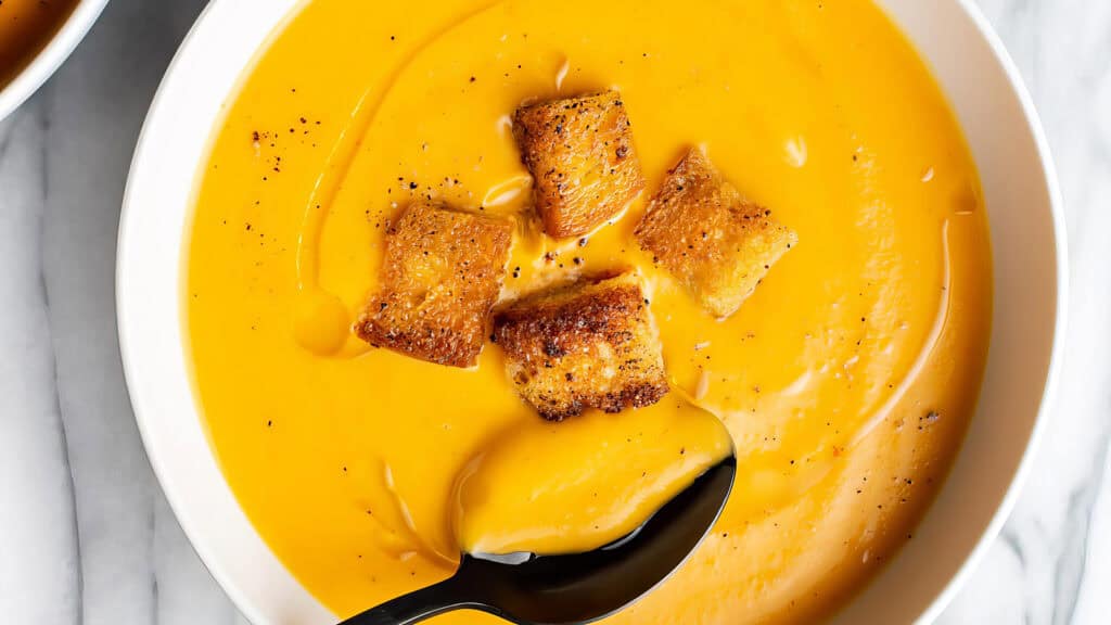 A bowl of creamy orange soup, inspired by Southern recipes, is garnished with four golden-brown croutons. A spoon rests partially submerged in the smooth, slightly glossy surface. The bowl is set on a pristine white marble surface.