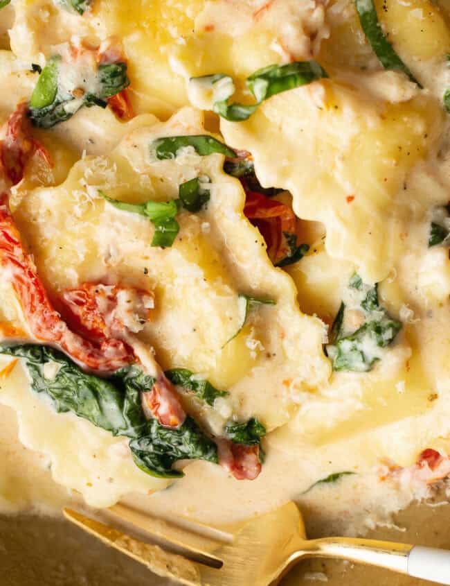 A close-up of a bowl of creamy ravioli, perfect for Pasta Lovers, garnished with shredded spinach and sun-dried tomatoes. The stuffed pasta is topped with a rich white sauce and grated cheese. A gold and white fork rests on the side of the bowl.