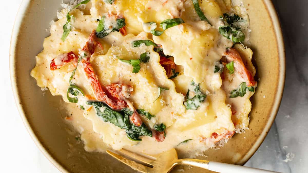 A close-up of a bowl of creamy ravioli, perfect for Pasta Lovers, garnished with shredded spinach and sun-dried tomatoes. The stuffed pasta is topped with a rich white sauce and grated cheese. A gold and white fork rests on the side of the bowl.