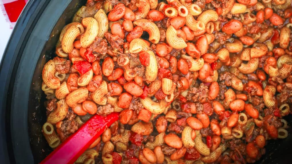 A close-up of a pot filled with a hearty dish combining elbow macaroni, ground beef, kidney beans, and diced tomatoes. The ingredients are well-mixed, coated in a rich tomato-based sauce perfect for ground beef recipes, and there's a red spoon stirring the mixture.