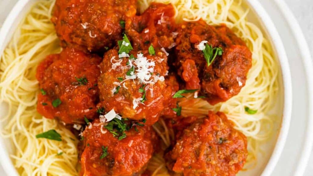 A bowl of spaghetti topped with several large meatballs in tomato sauce, one of the many delicious ground beef recipes, is garnished with grated parmesan cheese and chopped parsley. The dish is served on a white plate, highlighting the vibrant colors of the red sauce and green parsley.