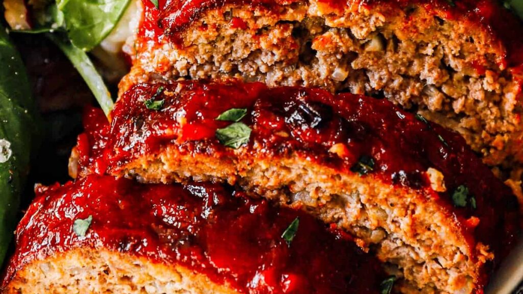 Close-up image of three slices of cooked meatloaf topped with a glossy red tomato-based glaze and garnished with chopped parsley. This savory ground beef recipe features a texture that appears moist and crumbly, with a side of leafy greens partially visible in the background.