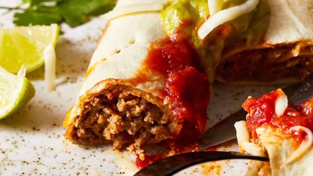 A close-up of Beef Taquitos filled with seasoned ground beef, topped with salsa, melted cheese, and guacamole. Part of the burrito is cut, revealing the filling. There are lime wedges and a leafy green garnish on the side. A fork and knife are also visible on the plate—perfect for those seeking ground beef recipes.