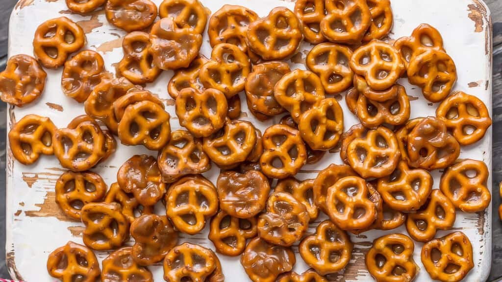 A close-up of numerous small pretzels covered in a glossy caramel coating, spread irregularly on a white, weathered wooden surface. These snack recipe favorites are shiny and golden brown, filling the frame with their distinct twisted shapes.