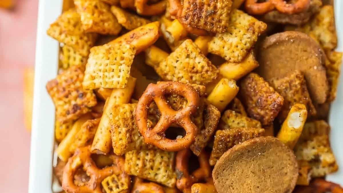 A close-up view of a snack mix featuring seasoned Chex cereal, pretzel twists, breadsticks, and round crackers, all coated with a savory seasoning. Perfect for any Snack Attack, the mix is displayed in a white serving tray. Delight Your Taste Buds with this fun and easy treat!