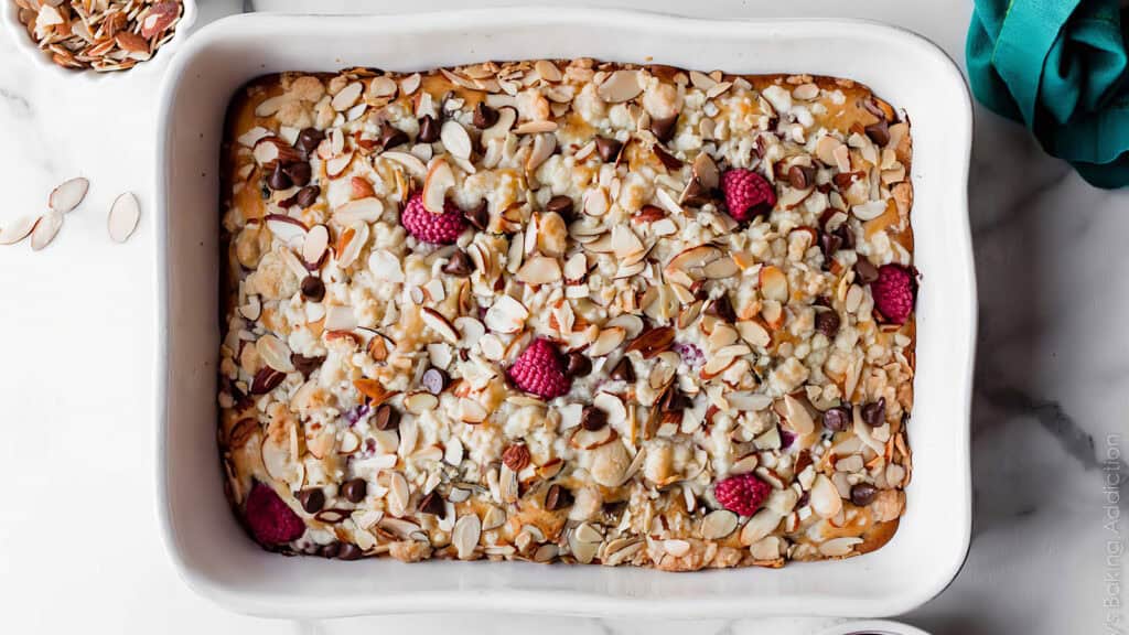 A white rectangular baking dish filled with a baked dessert topped with sliced almonds, chocolate chips, and fresh raspberries—a delightful take on sheet cake recipes. A small bowl of sliced almonds and a teal cloth add a charming touch in the background.