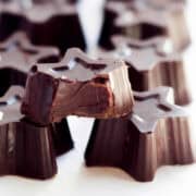 A close-up of star-shaped dark chocolate pieces arranged in rows. One piece is prominently in the foreground with a bite taken out of it, revealing the creamy interior. The background is blurred and white, resembling an enticing photo from gourmet snack recipes.