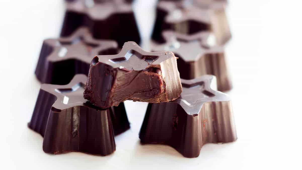 A close-up of star-shaped dark chocolate pieces arranged in rows. One piece is prominently in the foreground with a bite taken out of it, revealing the creamy interior. The background is blurred and white, resembling an enticing photo from gourmet snack recipes.