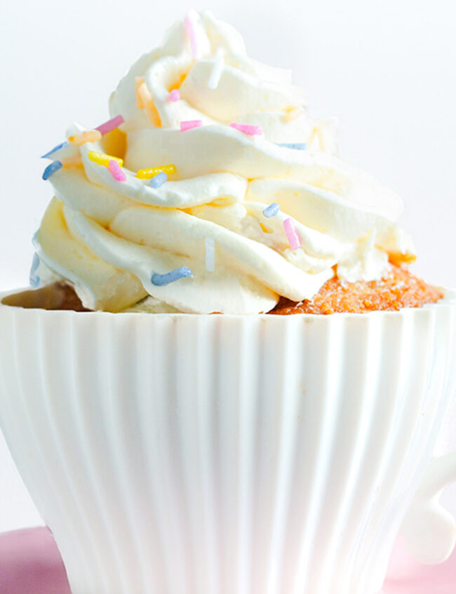 A white, ribbed teacup filled with a dessert topped with a generous swirl of frosting and colorful sprinkles. The teacup sits on a pink saucer against a plain white background, making it look like a mini version of cakes from beloved recipes.