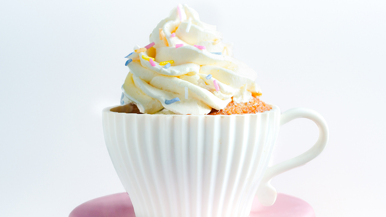 A white, ribbed teacup filled with a dessert topped with a generous swirl of frosting and colorful sprinkles. The teacup sits on a pink saucer against a plain white background, making it look like a mini version of cakes from beloved recipes.