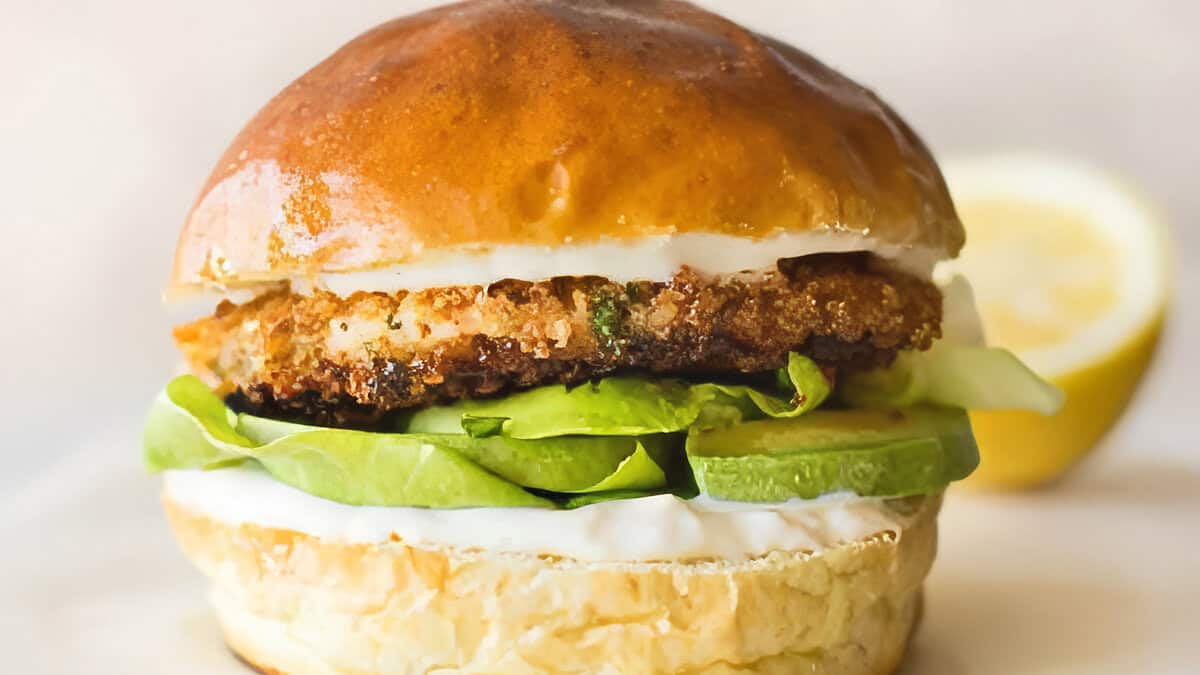 A close-up of a golden and crispy chicken burger with a glossy brioche bun. The burger is layered with fried chicken, sliced avocado, lettuce, and white sauce. A lemon wedge is partially visible in the background.