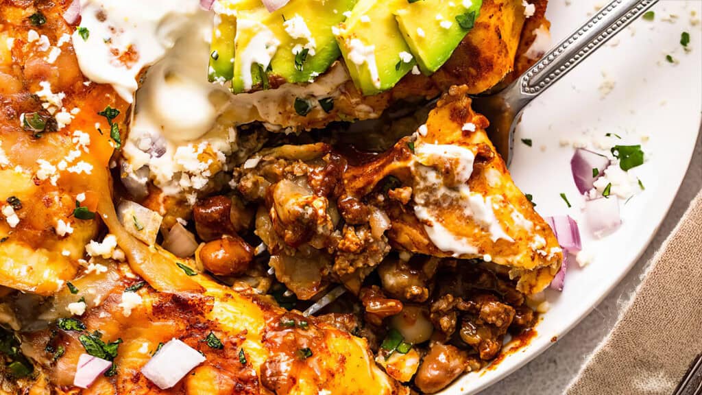 A close-up of a plate of enchiladas topped with cheese, sour cream, diced red onions, and avocado slices. The enchiladas are garnished with chopped cilantro and are filled with a savory mixture of beans and meat—perfect for lovers of ground beef recipes. A fork is placed beside the food.